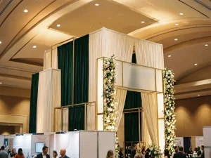 Hoog eventstand met bloemkolommen, hangende stoffen en verlichte kaders in ivoor, smaragdgroen en goud in een tentoonstellingshal.